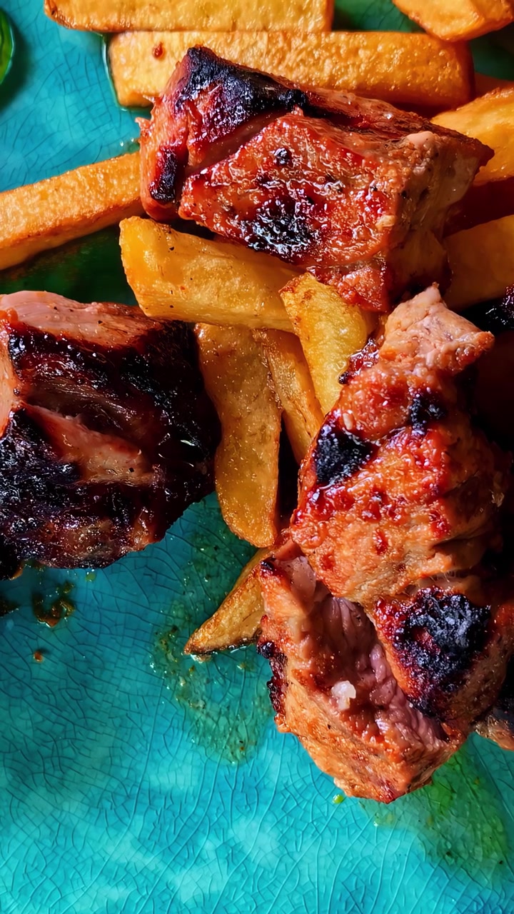 Primo piano di pezzi di carne grigliata carbonizzata abbinati a patatine fritte dorate su un piatto blu testurizzato. Ideale per blog di cucina o presentazioni culinarie.