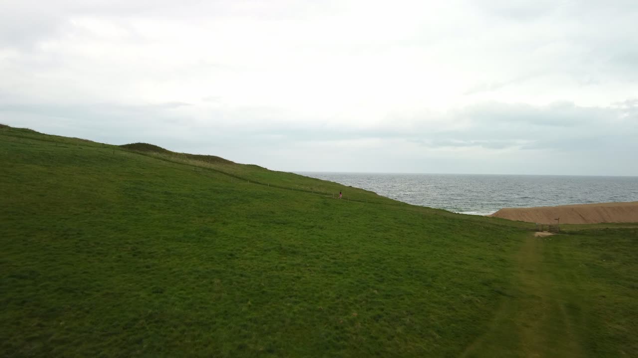 Aerial footage of a women walking down a grassy hill next to the sea on a cloudy day in Dorset on the Jurassic Coast. The footage moves straight forward with the women walking from left to right.