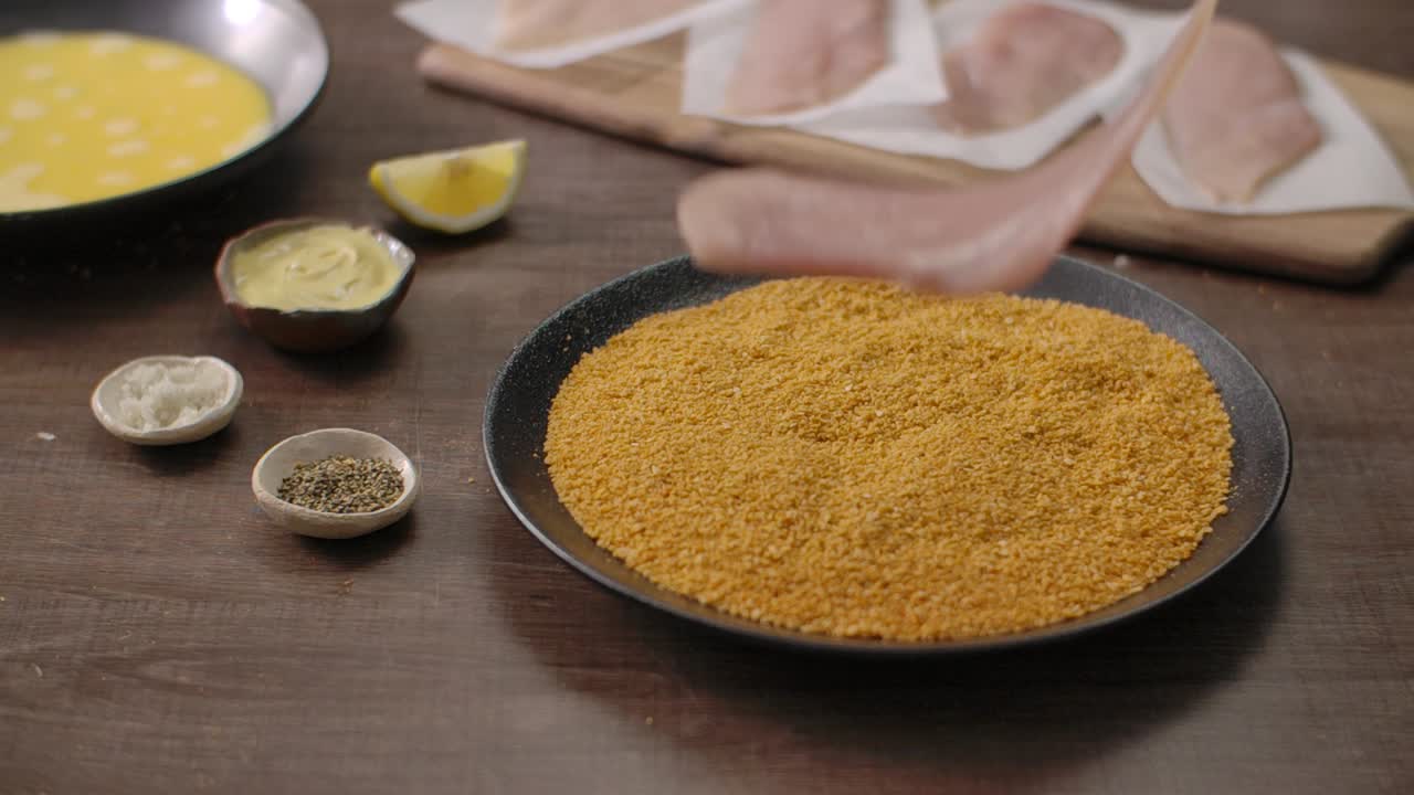 Close up shot of a fresh raw chicken fillet being placed on a plate of golden breadcrumbs for breading preparation in a kitchen