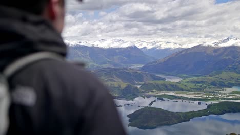 hombre mirando un hermoso valle verde con montañas sobre un lago en un día soleado en una colina cubierta de hierba, tiro de mano a la deriva