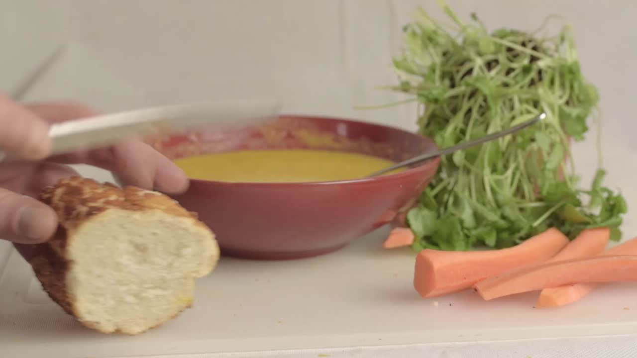 Hand cutting crusty bread with fresh carrot and coriander soup wide shot