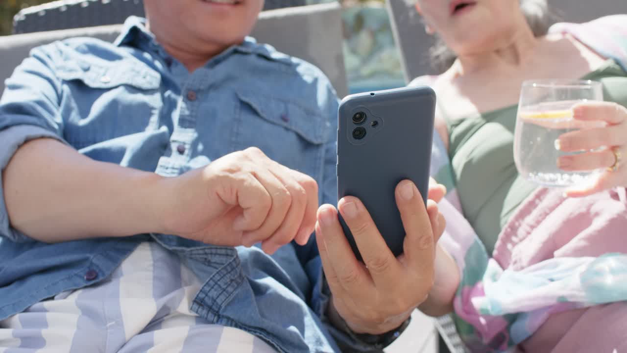 Diverse senior couple resting poolside, man tapping phone showing screen to woman with lemon glass
