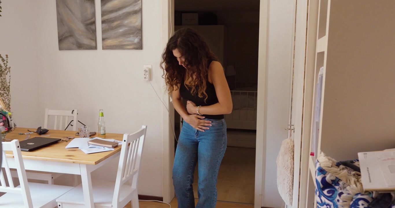 A woman bends over and holds her abdomen while experiencing sudden sharp abdominal pain at home.