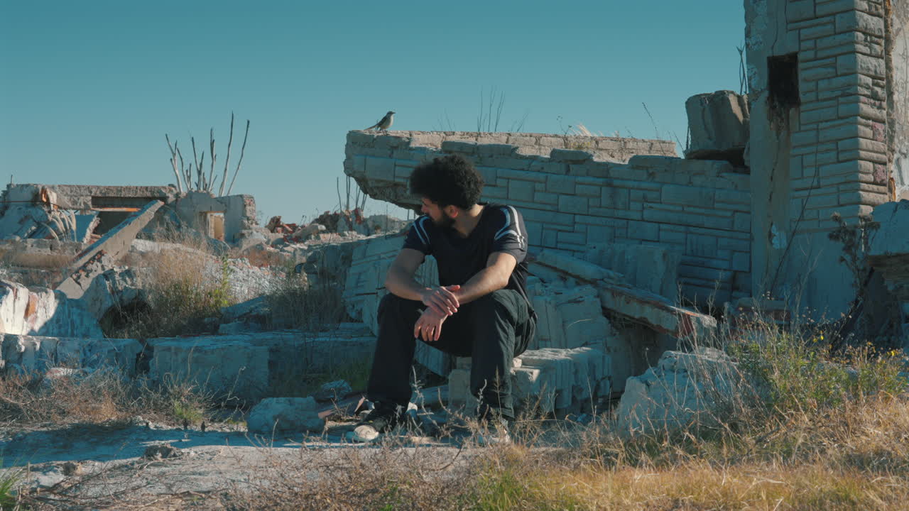 A thoughtful man sits quietly among collapsed stone structures and dry grass in a destroyed urban environment, expressing sadness, isolation, and reflection on loss and resilience