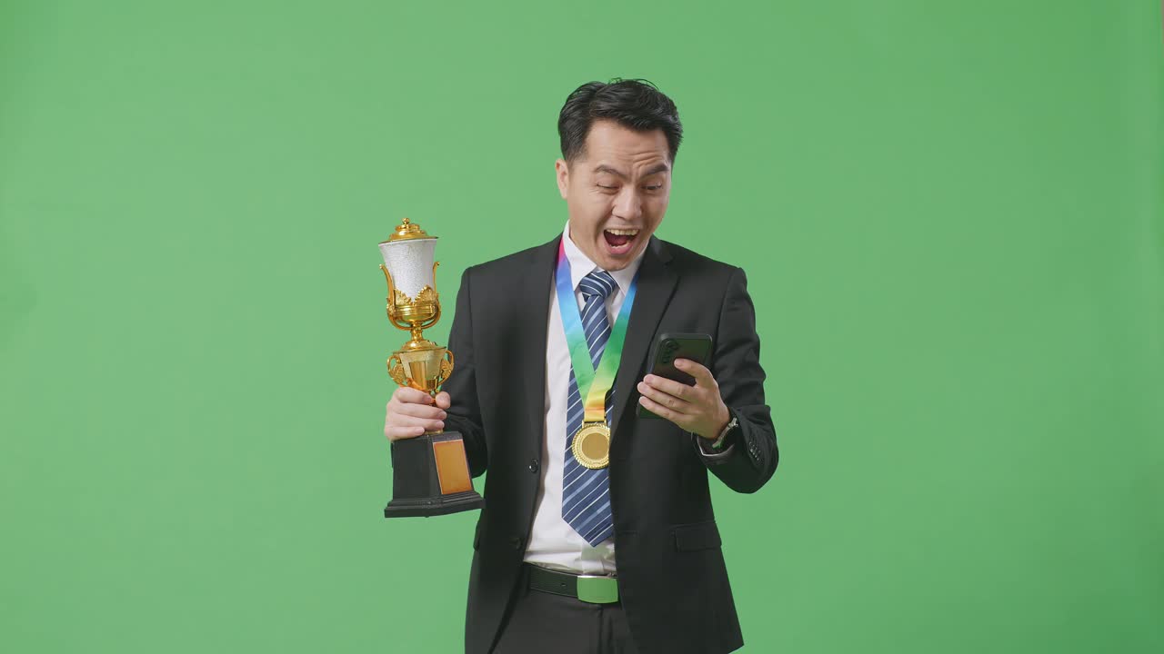 슈트와 타이를 입은 아시아 사업가, 금메달과 트로피를 들고 스마트폰을 보고 스튜디오의 녹색 화면 배경에서 와우라고 말합니다.