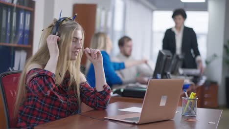 absorbed hipster woman listening to music in headphones and surfing internet on laptop with blurred colleagues discussing startup at background. young caucasian millennial working in office.