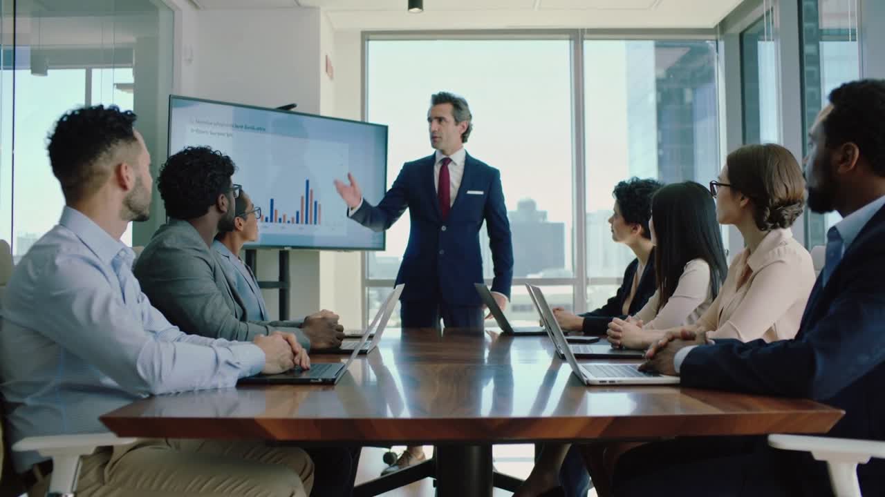 Pointing presenter suit red tie explaining bar chart at meeting start in meeting room with monitor
