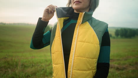 middle aged woman in green and black suit unzips her yellow jacket while standing in a vast open field under cloudy skies, with green trees visible in the distance