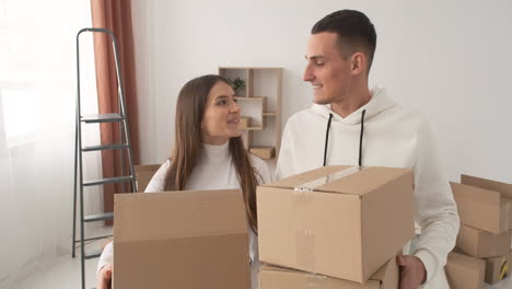 happy couple moving to new place holding cardboard boxes and walking together in empty flat 1