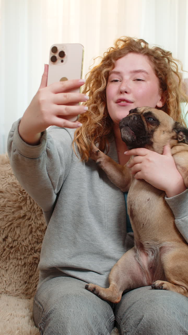 Young woman waving at video call on smartphone with pug dog on home sofa joyful friendly greeting