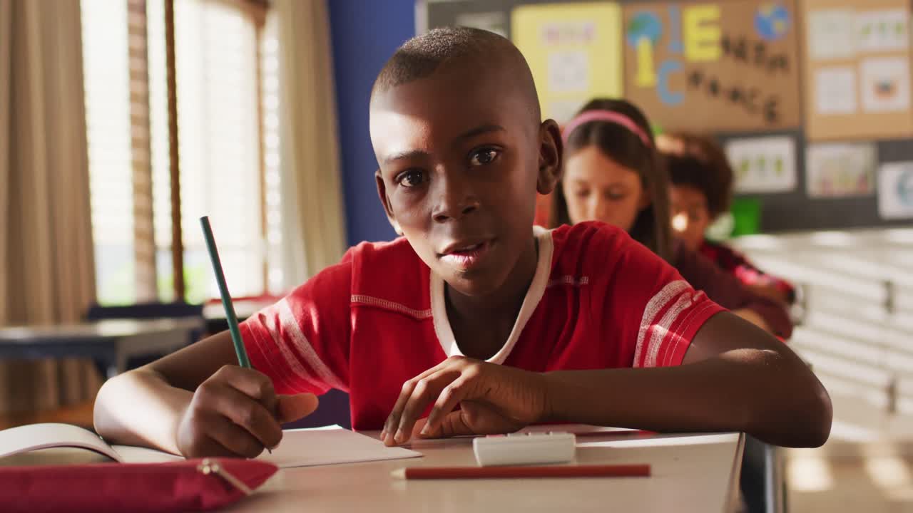 Portrait of african american schoolboy sitting in class, making notes, looking at camera
