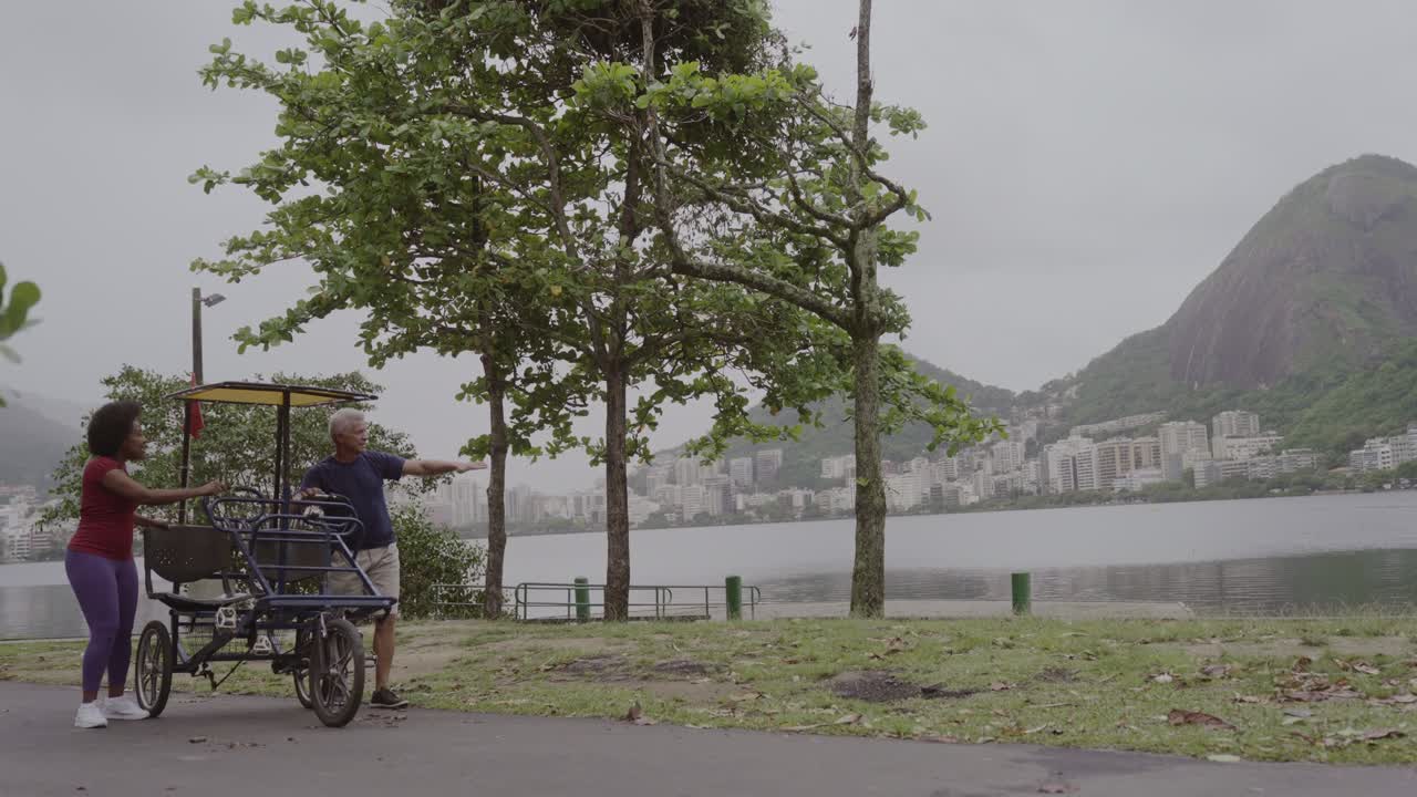 Two people with a pedal bike by a scenic lake with city and mountains in the background