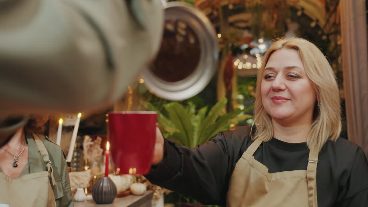 Due fioriste condividono caffè in serra. Fiorista bionda riceve tazza rossa versata dalla collega. Negozio caldo illuminato da candele, pieno di piante e decorazioni rustiche, pasticcini sul tavolo, conversazione rilassata.