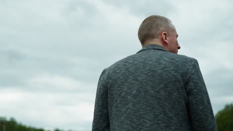 a close-up body view of an aged man wearing a gray jacket, seen from behind, with a sky in view