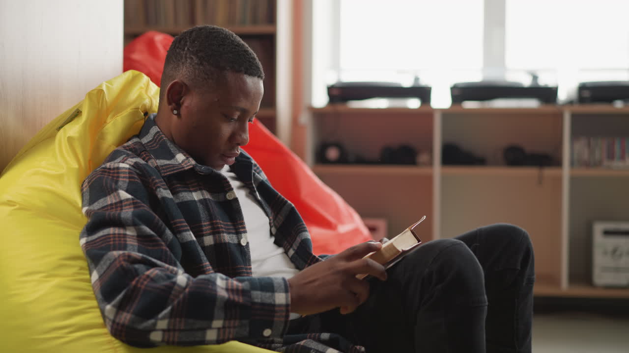 Young man reads book sitting on bean bag. Reader nestled in cushioned chair in students library lounge immersed in gripping novella. Comfort reading concept