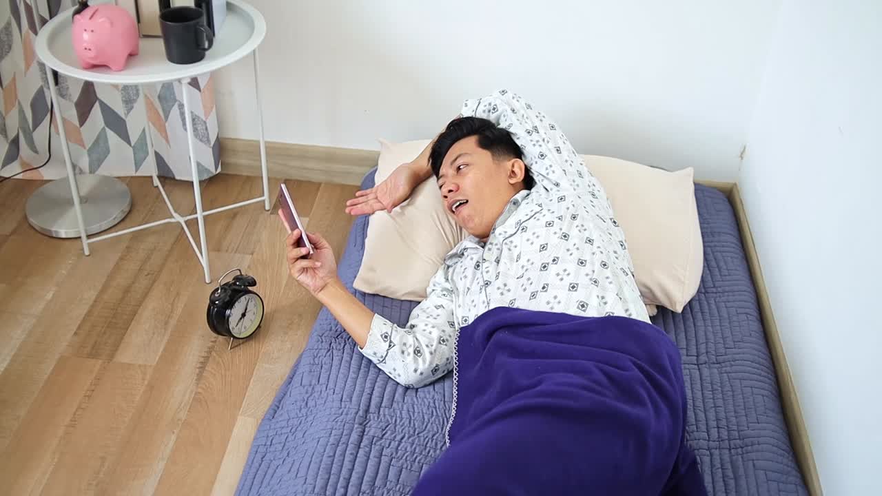 Young man happily scrolling phone while relaxing in bed with cozy blanket bright morning light