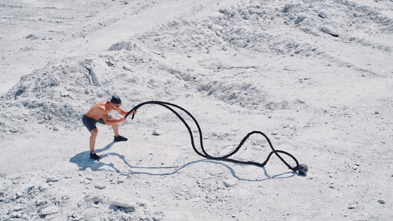 Strong young man doing exercises with a battle rope in sunny day. View from above on a muscular man training with cable on white ground outside. Concept of professional training.