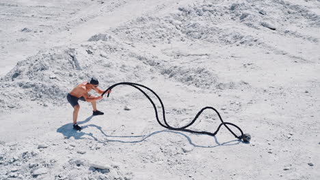 strong young man doing exercises with a battle rope in sunny day. view from above on a muscular man training with cable on white ground outside. concept of professional training.