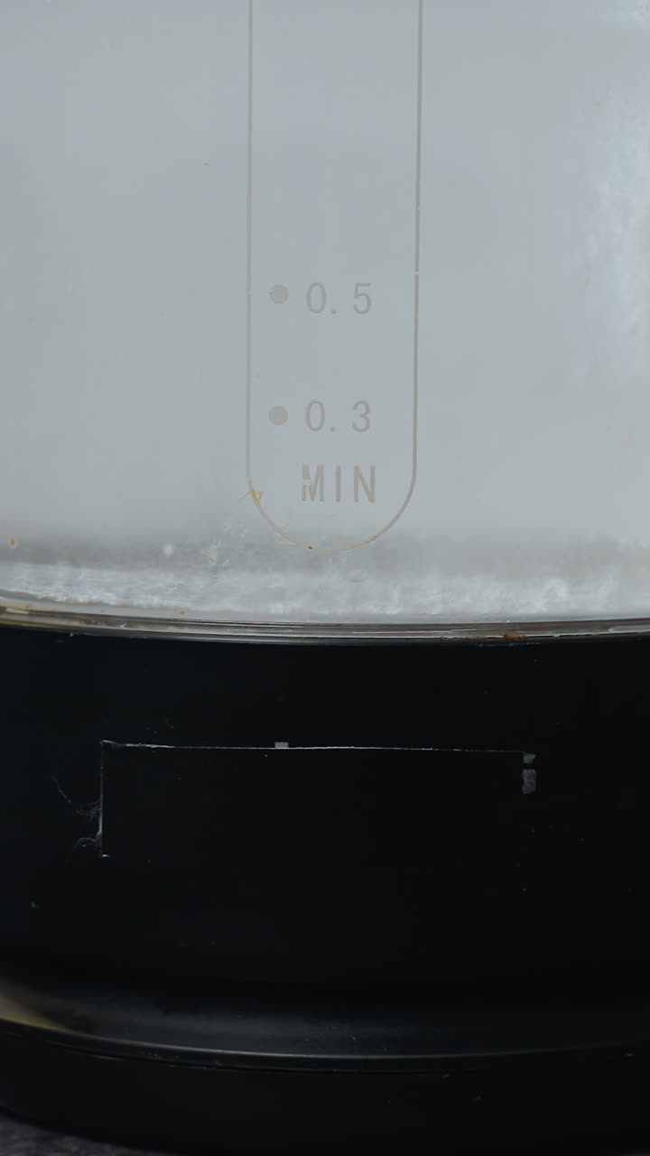 표면에 물 보일러를 놓고 있는 사람의 클로즈업, 물 수준과 항아리에 새겨진 글자에 초점을 맞추고, 가전제품과 쿠키 트레이가 있는 부의 일부를 볼 수 있습니다.