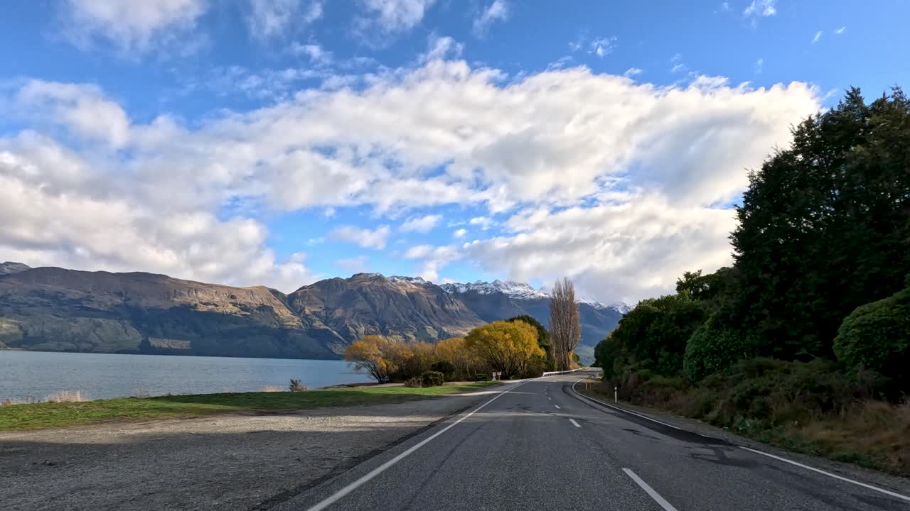 Car travels lakeside road with mountain views, autumn trees, dynamic clouds, and natural daylight