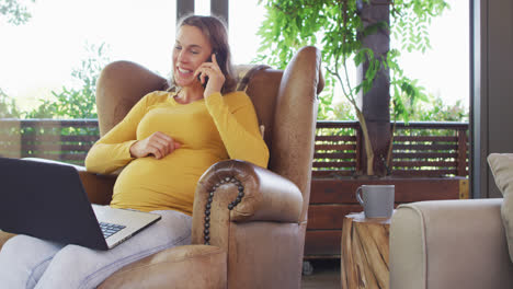 happy caucasian pregnant woman sitting in armchair having phone call and using laptop