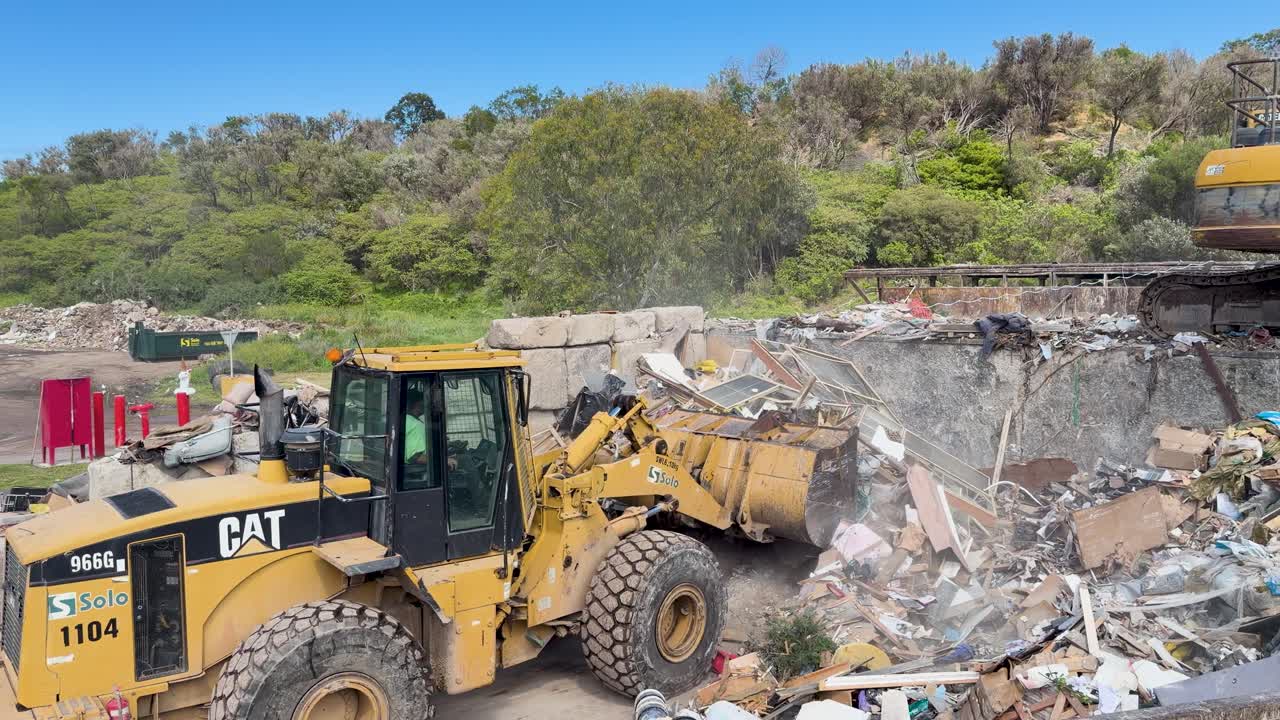Yellow front loader lifts and moves construction waste in bright daylight at outdoor landfill site