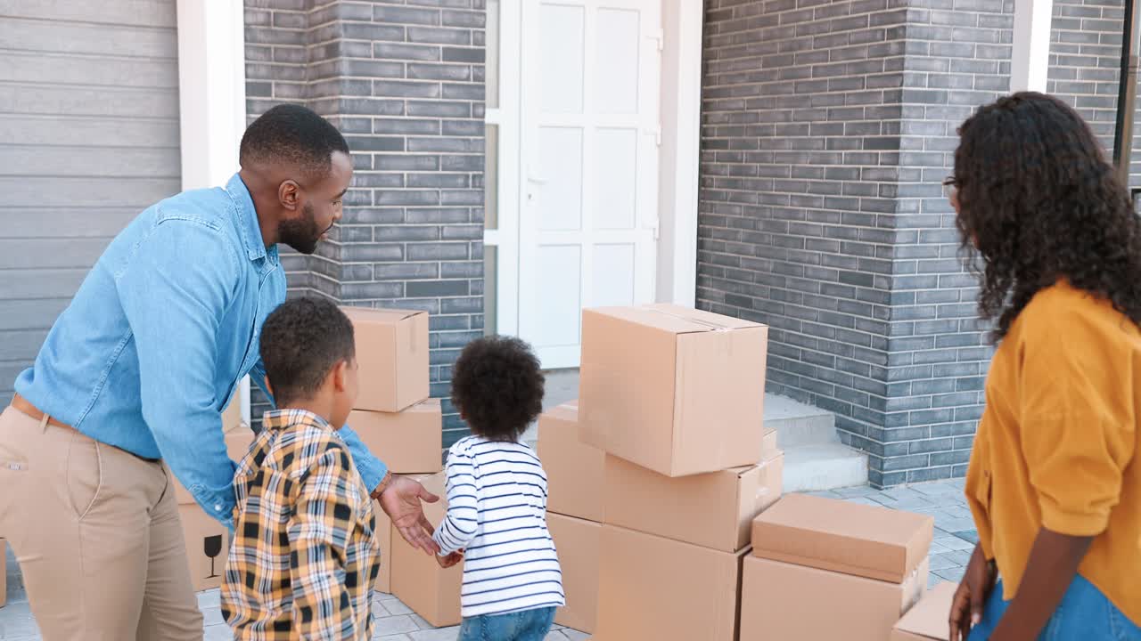 Happy African American parents with little children bringing carton boxes at yard of new home