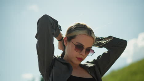 mujer con una camiseta negra debajo de una chaqueta gris, ajustando su cabello con una expresión enfocada bajo un cielo soleado y brillante, lleva gafas de sol, está al aire libre