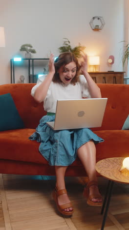 excited woman sitting on sofa using laptop receives good news victory celebrate lottery win at home