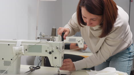 professional garment designer loosens presser foot of electric sewing machine with screwdriver while another designer in background works on fabric using identical machine in shared workshop setting