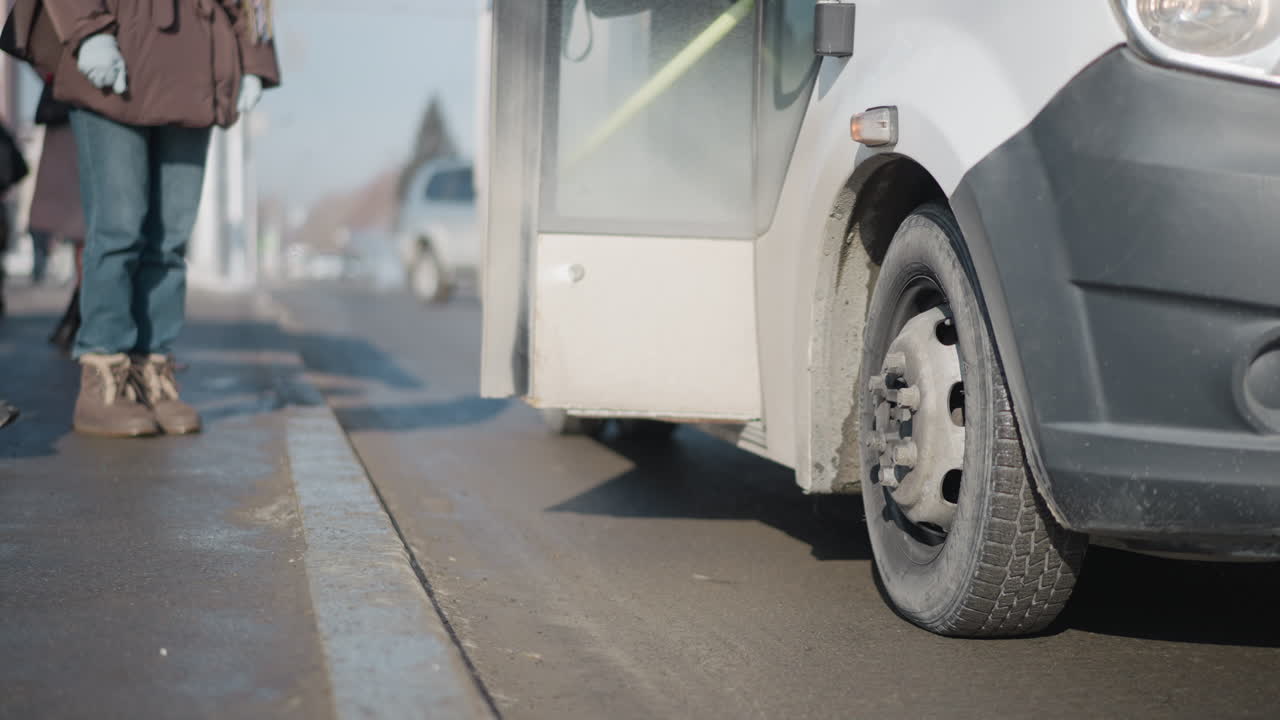 lower view shows passengers stepping out from bus at stop, boots landing on curb beside wheel, door open, winter light on asphalt and faint road mist, urban commute moment with motion