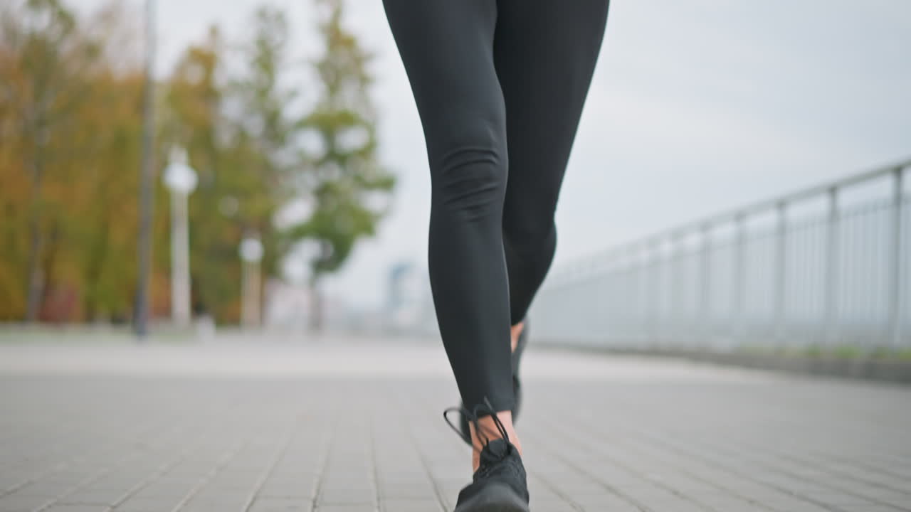 Close-up of woman s legs in black leggings and sneakers jogging in outdoor path with blurred background of trees, poles, and iron fence, showcasing fitness and active lifestyle