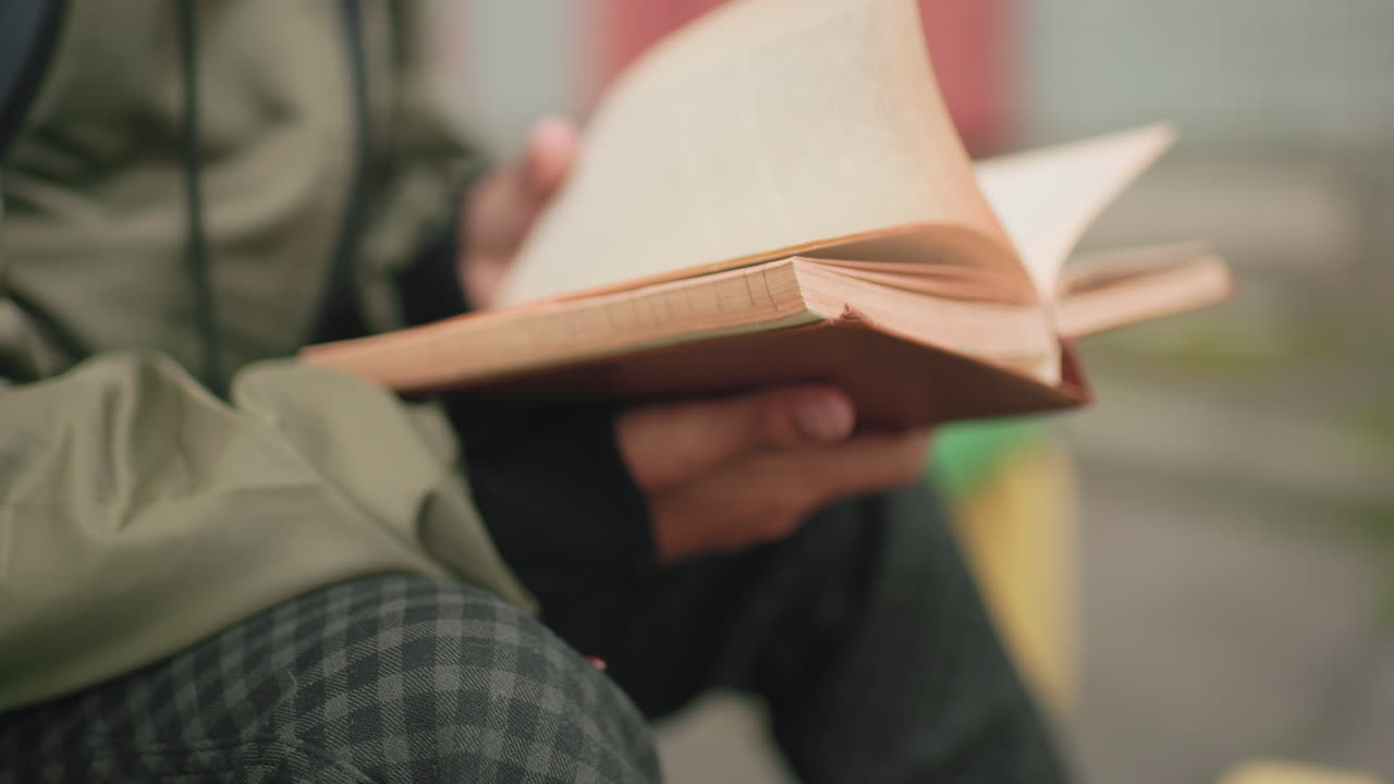 Close up side view of young kid wearing green jacket studying with focus while flipping to new page of book outdoors showing learning habit, and dedication to education with blurred background