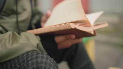 close up side view of young kid wearing green jacket studying with focus while flipping to new page of book outdoors showing learning habit, and dedication to education with blurred background