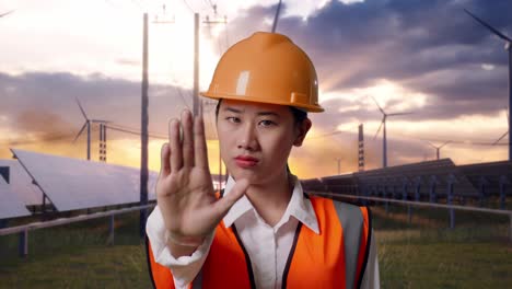 close up of asian female engineer with safety helmet disapproving with no hand sign with solar panel and wind turbines