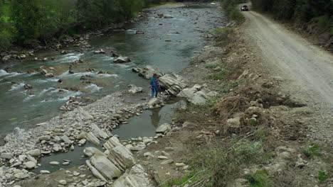 Woman-walking-rocky-river-shore-exploring-among-green-spruce-forest-enjoying