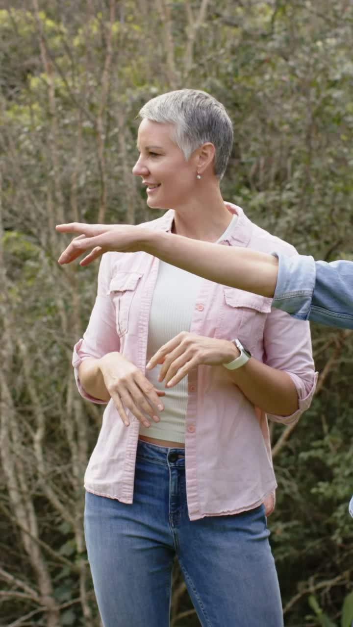 Video vertical: Mujer con top rosa girando, siguiendo un brazo de mezclilla desde la derecha que señala en el bosque para mirar