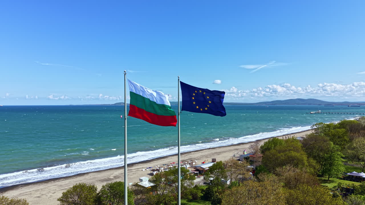 Bandiere della Bulgaria e dell'Unione Europea sventolano al vento sulla spiaggia in una giornata di sole, vicino a una località costiera con un cielo azzurro e limpido.