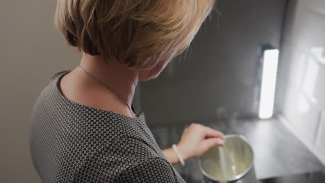 side view of female baker with short bob hairstyle wearing necklace and bracelet as she gently stirs bowl content using left hand and wooden stick in modern kitchen