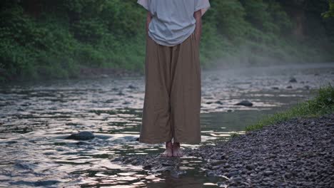 woman stands barefoot at the edge of a flowing river, embracing the tranquility of a serene morning, surrounded by mist and lush greenery