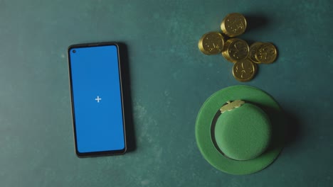 overhead studio shot of green leprechaun top hat with gold coins and blue screen mobile phone to celebrate st patricks day