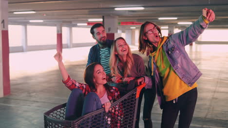 grupo joven de alegres amigos caucásicos divirtiéndose en un estacionamiento vacío con un carrito de supermercado y tomando selfies