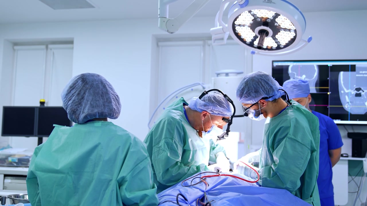 To kirurger læner sig over patienten under den skarpe lampe. En kvindelig sygeplejerske står ved siden af. En anæstesiolog går i baggrunden.