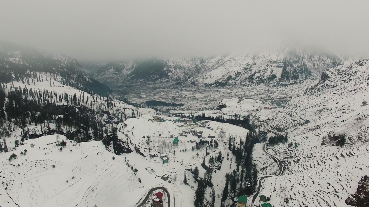 Aerial drone orbits over Gulaba Valley near Manali during active snowfall. Snow-covered mountain slopes, coniferous forests, winding road, and valley settlements visible under overcast winter sky.