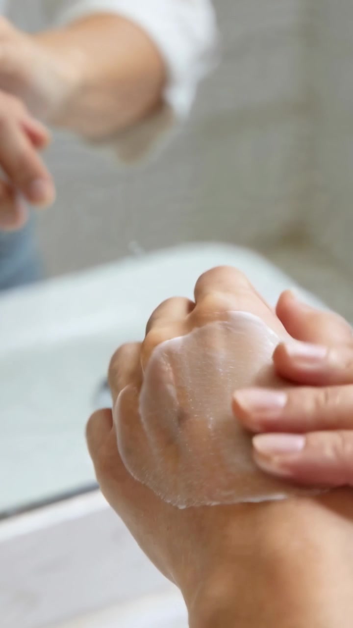 Video verticale: Donna che applica crema sulla mano, sfregando e idratando al lavandino, con le maniche bianche arrotolate.