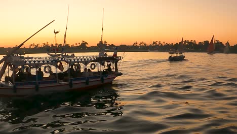 river scape of tourists sitting on egyption local felucca tour boats on nile river crossing from west to east bank with sunset colors reflecting on water. luxor, egypt