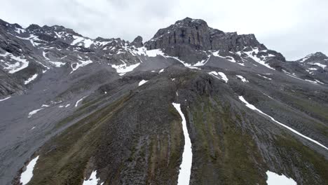 imágenes aéreas de drones que vuelan lentamente hacia arriba a través de un espectacular paisaje montañoso irregular, mirando hacia un imponente pico de montaña rocosa gris con parches residuales de nieve