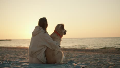 rear view of a blonde girl who sits next to her dog and interacts with her against the backdrop of the sea on a sunny morning