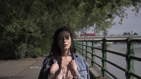 retrato de una atractiva turista latina con cabello negro ondulado y una chaqueta vaquera caminando por un parque en londres con vistas al puente putney detrás de ella