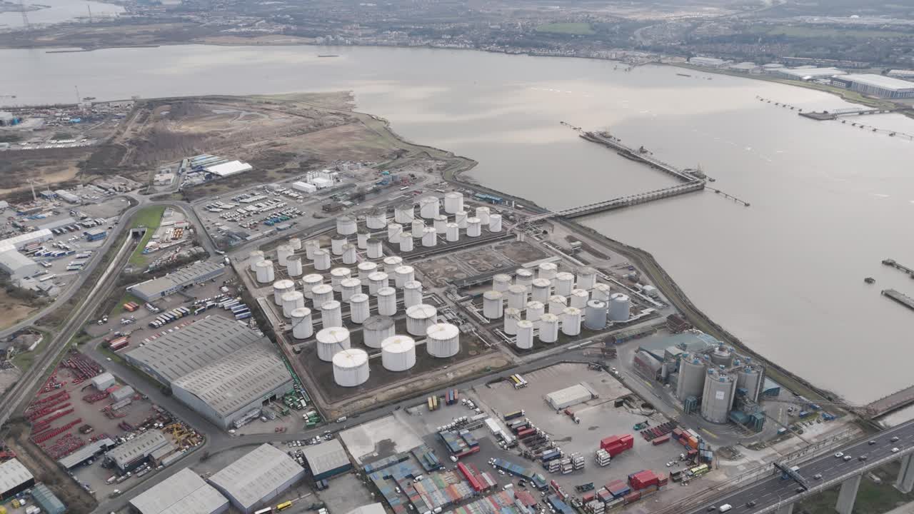 Aerial perspective of vast riverside tank farm with orderly rows of white cylindrical fuel silos, service roads and support structures beside the River Thames